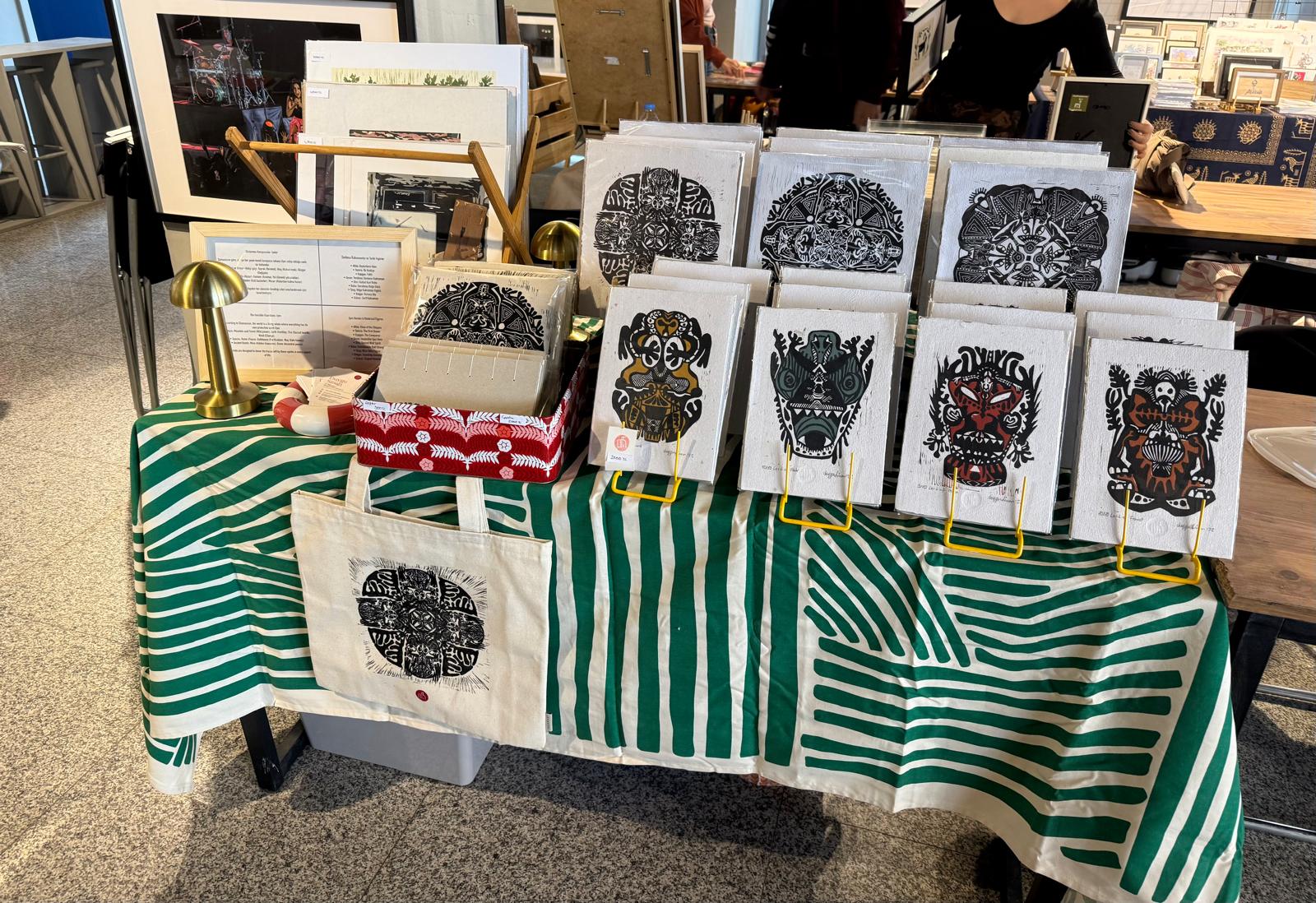 Maiamari standı UNITE'da — yeşil-beyaz örtü üzerinde linol baskı edisyonları ve tote bag